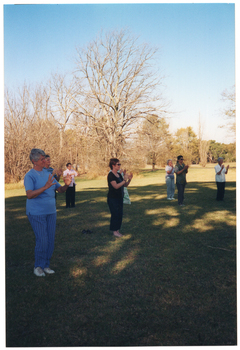 COL photo of a group of people standing on a grassy area in a large park. They are standing slightly apart, with their hands up in front of their chests. There are many trees in the background.