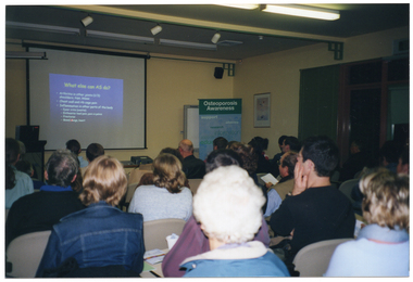 COL photo of a group of people seated on rows of chairs in a large room. They are watching slides which are being projected onto a large screen on the wall. Beside the projector screen is a freestanding, vertical, blue and green banner with the heading, "Osteoporosis Awareness", followed by more text, including the words "support", "advocacy", "research", "information", and "education".
