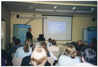 COL photo of a group of people in a large room. One man is standing next to a large slide projector screen at the top of the room. He is facing a group of people seated on rows of chairs. The slide being projected is titled, "What else can AS do?" On either side the projector screen, there is a large freestanding vertical banner: one banner is tiled, "Arthritis Victoria" and the other is titled, "Osteoporosis Awareness". Each banner contains words below the headings, such as "information", "support", "research", "advocacy", and "education".