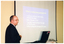 COL photo of a man standing next to a slide projector screen on the wall. The slide being projected is titled, "what is ankylosing spondylitis?" Below the heading, there is the following text on the slide: "A form of arthritis that mainly affects the spine"; "Male - Female = 2-3:1"; "Typically begins between the ages of 15-40 years"; and "Delay in diagnosis averages 5-7 years".