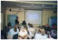 COL photo of a group of people in a large room. One man is standing next to a large slide projector screen at the top of the room. He is facing a group of people seated on rows of chairs. The slide being projected is titled, "What else can AS do?" On either side the projector screen, there is a large freestanding vertical banner: one banner is tiled, "Arthritis Victoria" and the other is titled, "Osteoporosis Awareness". Each banner contains words below the headings, such as "information", "support", "research", "advocacy", and "education".