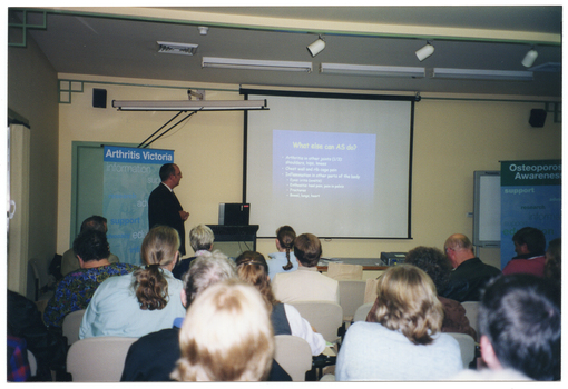 COL photo of a group of people in a large room. One man is standing next to a large slide projector screen at the top of the room. He is facing a group of people seated on rows of chairs. The slide being projected is titled, "What else can AS do?" On either side the projector screen, there is a large freestanding vertical banner: one banner is tiled, "Arthritis Victoria" and the other is titled, "Osteoporosis Awareness". Each banner contains words below the headings, such as "information", "support", "research", "advocacy", and "education".