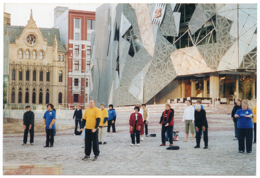 COL photo of a group of people standing in a paved town square. They are holding their arms straight, with flat palms, down in front of their thighs. One person has their arms up, palms downwards, at chest level. Behind them (in the right half of the frame), there are several wide corner steps leading to a large modern building. At the top of the building, there is an "SBS" television network logo. In the distance, to the left of the frame, there is a cathedral and a multistory building.
