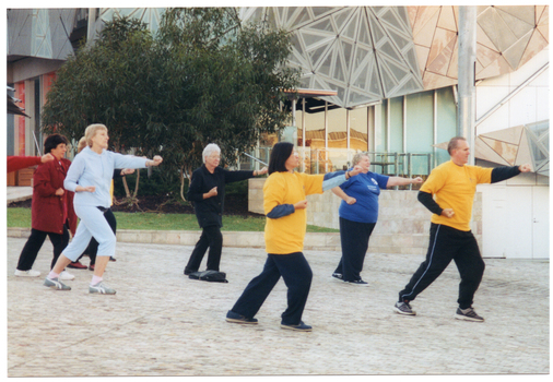 COL photo of a group of people exercising in a paved town square. They are lunging forward, with one arm outstretched in front of them and the other arm positioned near the chest or hip. Both hands have been formed into fists. In the background, there is a building with some mirrored glass panels and a small veranda. Beside the building, there is a grassy area with some trees at the back.
