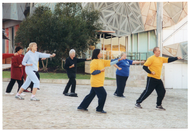 COL photo of a group of people exercising in a paved town square. They are lunging forward, with one arm outstretched in front of them and the other arm positioned near the chest or hip. Both hands have been formed into fists. In the background, there is a building with some mirrored glass panels and a small veranda. Beside the building, there is a grassy area with some trees at the back.