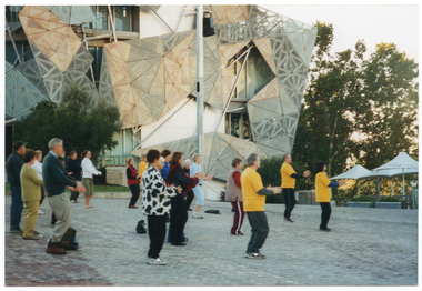 COL photo of a group of people exercising in a paved town square. They are standing with their arms slightly outstreched in from of them, at chest height. Some of the group are in shadow. In the background, there is a large yellow Edwardian building with a dome (partially obscured) on the roof.