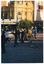 COL photo of a group of people exercising in a paved town square. They are standing with their arms slightly outstreched in from of them, at chest height. Some of the group are in shadow. In the background, there is a large yellow Edwardian building with a dome (partially obscured) on the roof.