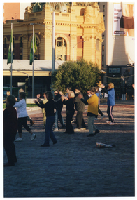 COL photo of a group of people exercising in a paved town square. They are standing with their arms slightly outstreched in from of them, at chest height. Some of the group are in shadow. In the background, there is a large yellow Edwardian building with a dome (partially obscured) on the roof.