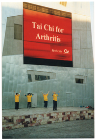 COL photo of four people standing beneath a large electronic advertising billboard on the side of a tall building. They are all wearing yellow t-shirts. The advertising billboard displays white text on a red background. The text is, "Tai Chi for Arthritis", with the respective text and logos for 'Arthritis Victoria' and 'Osteoporosis Victoria' below that.
