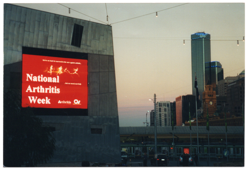 COL photo of a large electronic advertising billboard on the side of a tall building. The billboard displays white text on a red background. The text is, "National Arthritis Week", with the respective text and logos for 'Arthritis Victoria' and 'Osteoporosis Victoria' beside it. Above the main text are the silouhettes of some figures running and leaping. In the background, there is a view of some multistory buildings, including two tall skyscrapers.