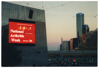COL photo of a large electronic advertising billboard on the side of a tall building. The billboard displays white text on a red background. The text is, "National Arthritis Week", with the respective text and logos for 'Arthritis Victoria' and 'Osteoporosis Victoria' beside it. Above the main text are the silouhettes of some figures running and leaping. In the background, there is a view of some multistory buildings, including two tall skyscrapers.