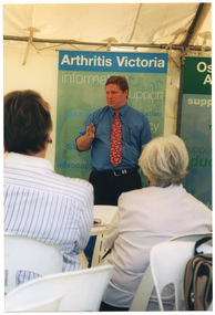 COL medium close up photo of a man standing in front of two freestanding vertical banners, speaking. In the foreground, there are two people seated on white plastic chairs at a white plastic table, facing him. Behind and above the banners, the internal metal frame of the white marquee in which they are located is evident. The headings on the banners are "Arthritis Victoria" and "Osteoporosis Awareness" (partially obscured). The text beneath the headings include words such as "information", "support", "research", "advocacy", and "education".