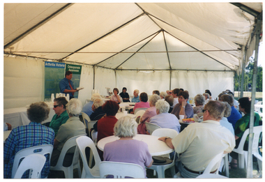COL wide frame, side view photo of a man standing in a large marquee, speaking to a group of people. He is looking down at some papers which he is holding in his hands. The group is sitting on white plastic chairs at a white plastic tables. On either side of the man speaking are two long rectangular tables covered with a white tablecloth. On top of the tables, there are glasses, polystyrene cups, and some printed materials such as brochures and/or books. Behind the man speaking, there are two freestanding vertical banners. The headings on the banners are "Arthritis Victoria" and "Osteoporosis Awareness". The text beneath the headings include words such as "information", "support", "research", "advocacy", and "education". The internal metal frame of the white marquee in which they are located is evident.
