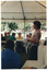 COL side view photo of a woman standing in a large marquee, speaking to a group of people. She is holding some papers in her hands. Beside her, there are several stacks of polystyrene cups on a table. The group is sitting on white plastic chairs at white plastic tables. The internal metal frame of the white marquee in which they are located is evident. In the background, there is a park with several large trees in the distance. There is a low level of light on the subjects inside the marquee, while the area outside (the park) is in full brightness.