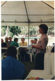 COL side view photo of a woman standing in a large marquee, speaking to a group of people. She is holding some papers in her hands. Beside her, there are several stacks of polystyrene cups on a table. The group is sitting on white plastic chairs at white plastic tables. The internal metal frame of the white marquee in which they are located is evident. In the background, there is a park with several large trees in the distance. There is a low level of light on the subjects inside the marquee, while the area outside (the park) is in full brightness.