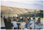 COL wide frame, side view photo of a man (partially obscured) standing in a large marquee, speaking to a group of people. He is in the background of the photo, at the edge of the left hand side of the frame. The group is sitting on white plastic chairs at a white plastic tables. In the foreground, on one side of the man speaking, there is a long rectangular table covered with a white tablecloth. On top of the table, at the far end, there are stacked glasses and polystyrene cups, and some paper napkins.  On top of the table, in the foreground, there are two stacks of black plasctic plates. The internal metal frame of the white marquee in which they are located is evident. In the background, outside of the marquee, there is a large park with several trees.