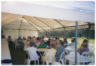COL wide frame, side view photo of a man (partially obscured) standing in a large marquee, speaking to a group of people. He is in the background of the photo, at the edge of the left hand side of the frame. The group is sitting on white plastic chairs at a white plastic tables. In the foreground, on one side of the man speaking, there is a long rectangular table covered with a white tablecloth. On top of the table, at the far end, there are stacked glasses and polystyrene cups, and some paper napkins.  On top of the table, in the foreground, there are two stacks of black plasctic plates. The internal metal frame of the white marquee in which they are located is evident. In the background, outside of the marquee, there is a large park with several trees.