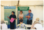 COL photo of three people standing inside a white marquee. One woman is speaking, while the other woman and a man are standing beside her with their arms crossed. They are facing a group of people who are sitting on white plastic chairs at white plastic tables. Behind them, there are two freestanding vertical banners. The headings on the banners are "Arthritis Victoria" and "Osteoporosis Awareness". The text beneath the headings include words such as "information", "support", "research", "advocacy", and "education". To the side of one banner, behind the man, stacks of polystyrene cups, and bowls containing tea bags and sugar satchets are evident at the far end of a table. The internal metal frame of the white marquee in which they are located is evident.
