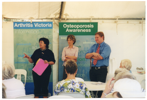COL photo of three people standing inside a white marquee. One woman is speaking, while the other woman and a man are standing beside her with their arms crossed. They are facing a group of people who are sitting on white plastic chairs at white plastic tables. Behind them, there are two freestanding vertical banners. The headings on the banners are "Arthritis Victoria" and "Osteoporosis Awareness". The text beneath the headings include words such as "information", "support", "research", "advocacy", and "education". To the side of one banner, behind the man, stacks of polystyrene cups, and bowls containing tea bags and sugar satchets are evident at the far end of a table. The internal metal frame of the white marquee in which they are located is evident.