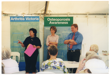 COL photo of three people standing inside a white marquee. One woman is speaking, while the other woman and a man are standing beside her with their arms crossed. They are facing a group of people who are sitting on white plastic chairs at white plastic tables. Behind them, there are two freestanding vertical banners. The headings on the banners are "Arthritis Victoria" and "Osteoporosis Awareness". The text beneath the headings include words such as "information", "support", "research", "advocacy", and "education". To the side of one banner, behind the man, stacks of polystyrene cups, and bowls containing tea bags and sugar satchets are evident at the far end of a table. The internal metal frame of the white marquee in which they are located is evident.