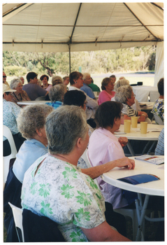 COL side view photo of a group of people sitting on white plastic chairs at white plastic tables. On various tables, there are some printed booklets and some plastic or paper tumblers. The internal metal frame of the white marquee in which they are located is evident. In the background, outside of the marquee, there is a large park with several trees.