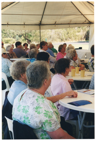 COL side view photo of a group of people sitting on white plastic chairs at white plastic tables. On various tables, there are some printed booklets and some plastic or paper tumblers. The internal metal frame of the white marquee in which they are located is evident. In the background, outside of the marquee, there is a large park with several trees.