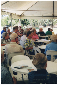 COL side view photo of a group of people sitting on white plastic chairs at white plastic tables. On various tables, there are some printed booklets and one polystyrene cup. The internal metal frame of the white marquee in which they are located is evident. In the background, outside of the marquee, there is a large park with several trees.