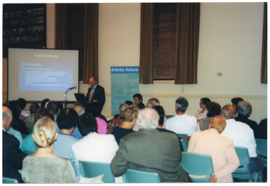 COL photo of a man, standing, addressing a roomful of people. He is speaking into a microphone, at a lectern, to an audience who are seated on several rows of chairs. Beside him is a projector screen showing a slide titled, "Types of arthritis". Behind him is a freestanding vertical blue and green banner with the heading, 'Arthritis Victoria'. Next to him is a table (partially obscured) at which three other people (partially obscured) are seated. They are all facing the audience who are in the foreground, and have been Photographed from behind.
