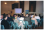COL photo of a woman, standing, addressing a roomful of people. She is speaking into a microphone, at a lectern, to an audience who are seated on several rows of chairs. Beside her is a projector screen showing a slide. Behind her is a freestanding vertical blue and green banner with the heading, 'Arthritis Victoria'. Next to her is a table (partially obscured) at which three other people (partially obscured) are seated. They are all facing the audience who are in the foreground, and have been Photographed from behind.