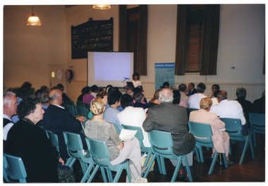 COL photo of a woman, standing, addressing a roomful of people. She is speaking into a microphone, at a lectern, to an audience who are seated on several rows of chairs. Beside her is a projector screen showing a slide. Behind her is a freestanding vertical blue and green banner with the heading, 'Arthritis Victoria'. Next to her is a table (partially obscured) at which three other people (partially obscured) are seated. They are all facing the audience who are in the foreground, and have been Photographed from behind.