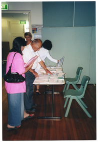 COL photo of three people perusing a range of information sheets which have been laid out on a long rectangular table. There are also some brochures on a stand on the table. Standing beside them, near a doorway, is a woman (partially obscured) who has one hand to her lips while looking down. Another room is evident through the doorway, with an "EXIT" sign above a second doorway.