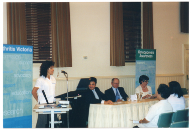 COL photo of a woman, standing, speaking into a microphone at a lectern. There is a small table in front of her, holding a slide projector and a few small items. Beside her, there are three people sitting at a long rectangular wooden table, around which there is a cream valance. Behind the woman and the people seated are two tall windows above a dado wall. The windows are dressed with dark wooden venetian blinds and brown curtains. Beside each window, there is a tall, freestanding, blue and green banner. The headings on the banners are, respectively, "Arthritis Victoria" and "Osteoporosis Awareness". The text beneath the headings include words such as "information", "support", "research", "advocacy", and "education". In the foreground, there are two audience members sitting in the front row.