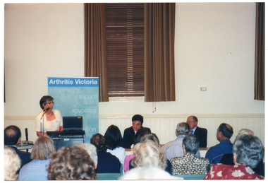COL photo of a woman, standing, addressing a roomful of people. She is speaking into a microphone, at a lectern, to an audience who are seated on several rows of chairs. Behind her is a freestanding vertical blue and green banner with the heading, 'Arthritis Victoria'. Next to her is a table (partially obscured) at which two men (partially obscured) are seated. They are all facing the seated audience who are in the foreground, and have been Photographed from behind.