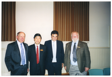 COL photo of four men, standing side by side, posing for a photo. Behind them are two tall windows, with closed brown curtains, above a dado wall. There are some chairs to the right of the frame.