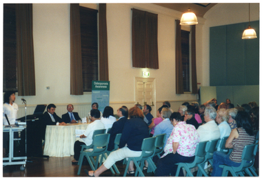 COL photo of a woman, standing, addressing a roomful of people. She is speaking into a microphone, at a lectern, to an audience who are seated on several rows of chairs. Beside her, there are three people sitting at a long rectangular wooden table, around which there is a cream valance. Behind the woman and the people seated are two tall windows above a dado wall. The windows are dressed with dark wooden venetian blinds and brown curtains. At the other end of the table is a freestanding vertical blue and green banner, titled 'Osteoporosis Awareness'. In the foreground, the audience is seated on several rows of chairs. They have been Photographed from behind at a slight angle.