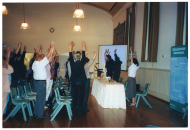 COL side view photo of a roomful of people in a hall. To the right of the frame, four people are standing behind a long rectangular table with a cream valance around it. They are facing an audience (centre and left of the frame) who are standing between rows of plastic chairs. Everyone has their arms outstretched above their heads. Behind the four people at the table, there are three tall windows above a dado wall and a large wooden honour board further back along the wall. There is a slide projector screen between the honour board and the table. There are two vertical freestanding blue and green banners along the wall behind the table. One banner is in the foreground, to the right of the frame. It contains the heading, "Osteoporosis Awareness", followed by words such as "information", "support", "research", "advocacy", and "education".