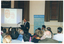 COL photo of a man, standing behind a lectern, addressing a roomful of people. He is speaking into a microphone to an seated audience. Beside him is a slide projector screen displaying a slide with some images. Behind him, there is a freestanding vertical blue and green banner with the heading, 'Arthritis Victoria'. Below the heading are words such as "information", "support", "research", "advocacy", and "education". On the wall behind him, there are two tall windows with wooden venetian blinds and brown curtains above a dado wall. On the wall beside one window, there is a large wooden honour board. Next to him is a table (partially obscured) at which a man (partially obscured) is seated. They are facing the audience who is in the foreground, and has been Photographed from behind.