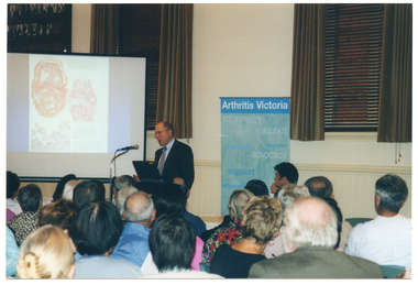 COL photo of a man, standing behind a lectern, addressing a roomful of people. He is speaking into a microphone to an seated audience. Beside him is a slide projector screen displaying a slide with some images. Behind him, there is a freestanding vertical blue and green banner with the heading, 'Arthritis Victoria'. Below the heading are words such as "information", "support", "research", "advocacy", and "education". On the wall behind him, there are two tall windows with wooden venetian blinds and brown curtains above a dado wall. On the wall beside one window, there is a large wooden honour board. Next to him is a table (partially obscured) at which a man (partially obscured) is seated. They are facing the audience who is in the foreground, and has been Photographed from behind.