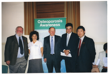 COL photo of five people standing side by side, posing for a photo. Behind them is a freestanding vertical blue and green banner, titled 'Osteoporosis Awareness'. Behind the banner is a tall window, with wooden venetian blinds and brown curtains, above a dado wall.  Behind them is a freestanding vertical blue and green banner, titled 'Osteoporosis Awareness'.