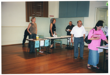 COL photo of two people standing behind some tables which are covered with information sheets and some leaflets on a stand. A man is standing beside the table, reading one of the brochures, which he is holding. In the foreground and to the right of the frame, a few people are perusing some information sheets and brochures on a table opposite. Further to the right, there is an  open doorway with an "EXIT" sign (partially obscured) above it.