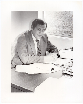 B&W photo of a man, sitting at a desk. He is wearing a shirt, suit and tie. He has his clasped hands resting on the desk, on which there are several small stacks of papers and a wire document tray. There is a window beside the desk, through which a grassy area is evident.