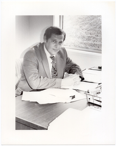 B&W photo of a man, sitting at a desk. He is wearing a shirt, suit and tie. He has his clasped hands resting on the desk, on which there are several small stacks of papers and a wire document tray. There is a window beside the desk, through which a grassy area is evident.