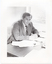 B&W photo of a man, sitting at a desk. He is wearing a shirt, suit and tie. He has his clasped hands resting on the desk, on which there are several small stacks of papers and a wire document tray. There is a window beside the desk, through which a grassy area is evident. The light exposure on this photo print is too high, thus providing low contrast.