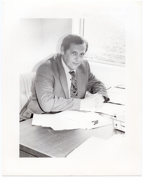 B&W photo of a man, sitting at a desk. He is wearing a shirt, suit and tie. He has his clasped hands resting on the desk, on which there are several small stacks of papers and a wire document tray. There is a window beside the desk, through which a grassy area is evident. The light exposure on this photo print is too high, thus providing low contrast.