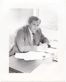 B&W photo of a man, sitting at a desk. He is wearing a shirt, suit and tie. He has his clasped hands resting on the desk, on which there are several small stacks of papers and a wire document tray. There is a window beside the desk, through which a grassy area is evident. The light exposure on this photo print is too high, thus providing low contrast.