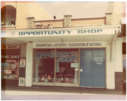 COL photo of the exterior of a shop, with two other shops (partially obscured) evident on either side. On the outside of the veranda, the RAAV logo and "OPPORTUNITY SHOP" are painted in blue onto a white background. Above the shop's display window and double doors (beneath the veranda), the text "RHEUMATISM & ARTHRITIS ASSOCIATION OF VICTORIA" is painted in blue on a white background. On the window glass, the words "OPPORTUNITY SHOP" are painted in blue, in a tapered design. The double front doors are also painted in the same blue, and on one door, the text "RHEUMATISM AND ARTHRITIS ASSOCIATION OF VICTORIA", "ACTION HOUSE YARRA BOULEVARD KEW 3101 Phone 862 2555" are painted in white. Below this, the numbers "1428" (the street number of the building) is also painted in white. Behind the window, various items are on display. Inside the window, near the doors, a handwritten paper sign has been affixed which says, "OPENING MONDAY 10:30". To the left of the shop, there is a newsagency (partially obscured). Above the window, and beneath the verandah, there is the text (partially obscured), "...e Sun" with a picture of a rooster between the two words. At the top of the window, there is a sign containing the text, "THE AGE". Two bill posters inside wire cages are affixed to the wall. One says, "The AGE", "LIBS DEFY FRASER". The other says, "WEEKEND AUSTRALIAN", "THREE MILLION TO DIE", and "THE LIFE OF BILLY HUGHES".