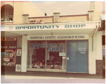 COL photo of the exterior of a shop, with two other shops (partially obscured) evident on either side. On the outside of the veranda, the RAAV logo and "OPPORTUNITY SHOP" are painted in blue onto a white background. Above the shop's display window and double doors (beneath the veranda), the text "RHEUMATISM & ARTHRITIS ASSOCIATION OF VICTORIA" is painted in blue on a white background. On the window glass, the words "OPPORTUNITY SHOP" are painted in blue, in a tapered design. The double front doors are also painted in the same blue, and on one door, the text "RHEUMATISM AND ARTHRITIS ASSOCIATION OF VICTORIA", "ACTION HOUSE YARRA BOULEVARD KEW 3101 Phone 862 2555" are painted in white. Below this, the numbers "1428" (the street number of the building) is also painted in white. Behind the window, various items are on display. Inside the window, near the doors, a handwritten paper sign has been affixed which says, "OPENING MONDAY 10:30". To the left of the shop, there is a newsagency (partially obscured). Above the window, and beneath the verandah, there is the text (partially obscured), "...e Sun" with a picture of a rooster between the two words. At the top of the window, there is a sign containing the text, "THE AGE". Two bill posters inside wire cages are affixed to the wall. One says, "The AGE", "LIBS DEFY FRASER". The other says, "WEEKEND AUSTRALIAN", "THREE MILLION TO DIE", and "THE LIFE OF BILLY HUGHES".