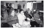 B&W photo of a small group of people sitting on rows of chairs in a room. One woman in the front row is looking directly at the camera. The three other women in the front row are holding cups and saucers - one of them is drinking from the cup while the other two are holding them in their laps. One man in the second row is turning around to face another man in the third row. In the background, there is a tall filing cabinet in the corner, near a large window. On the other side of the window, there is a large multipanel display board. There are a few pictures and/or posters and a large framed mirror on the walls.
