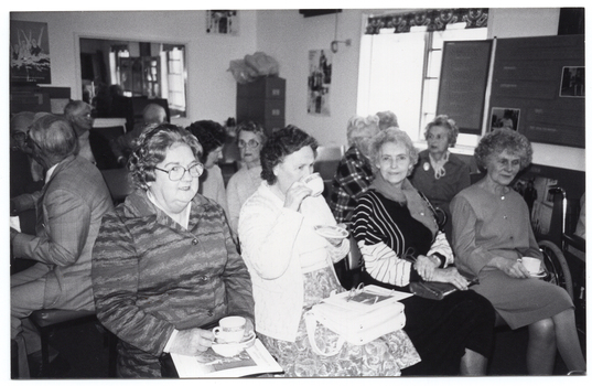 B&W photo of a small group of people sitting on rows of chairs in a room. One woman in the front row is looking directly at the camera. The three other women in the front row are holding cups and saucers - one of them is drinking from the cup while the other two are holding them in their laps. One man in the second row is turning around to face another man in the third row. In the background, there is a tall filing cabinet in the corner, near a large window. On the other side of the window, there is a large multipanel display board. There are a few pictures and/or posters and a large framed mirror on the walls.