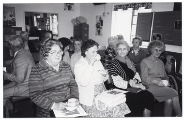 B&W photo of a small group of people sitting on rows of chairs in a room. One woman in the front row is looking directly at the camera. The three other women in the front row are holding cups and saucers - one of them is drinking from the cup while the other two are holding them in their laps. One man in the second row is turning around to face another man in the third row. In the background, there is a tall filing cabinet in the corner, near a large window. On the other side of the window, there is a large multipanel display board. There are a few pictures and/or posters and a large framed mirror on the walls.